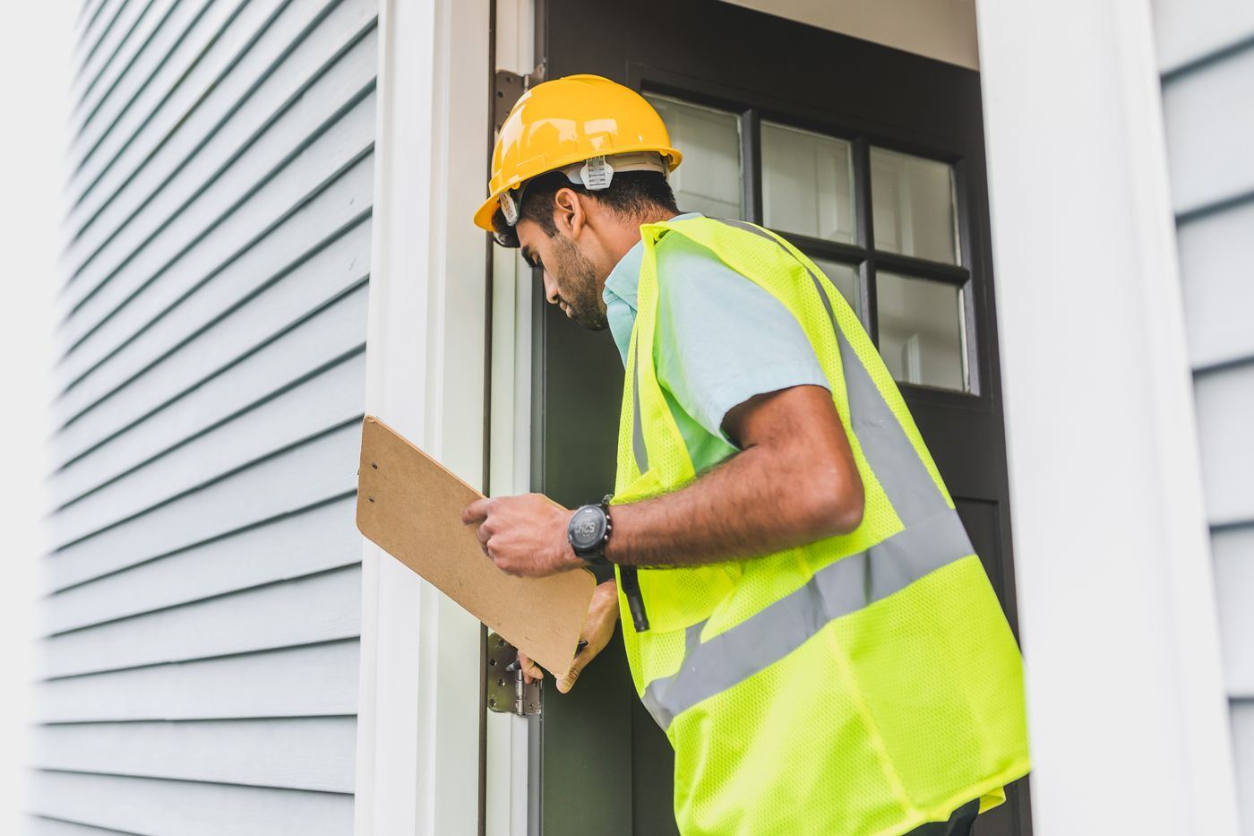 Surveyor in a hard hat and hi-vis vest inspecting a new home with a clipboard
